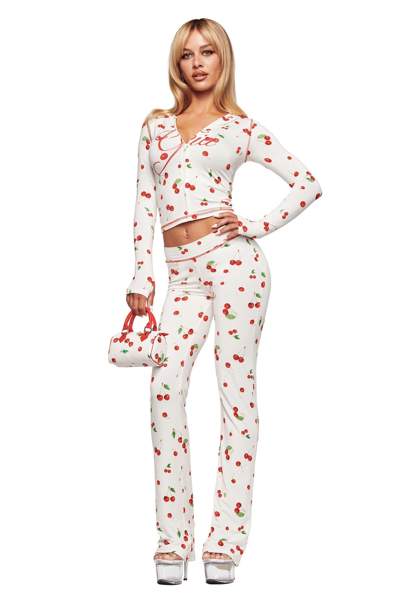 A woman wears the I.AM.GIA BLARE HOODIE in the Cherry on Top print, paired with matching pants and a small handbag, styled with silver platform heels for a bold look.