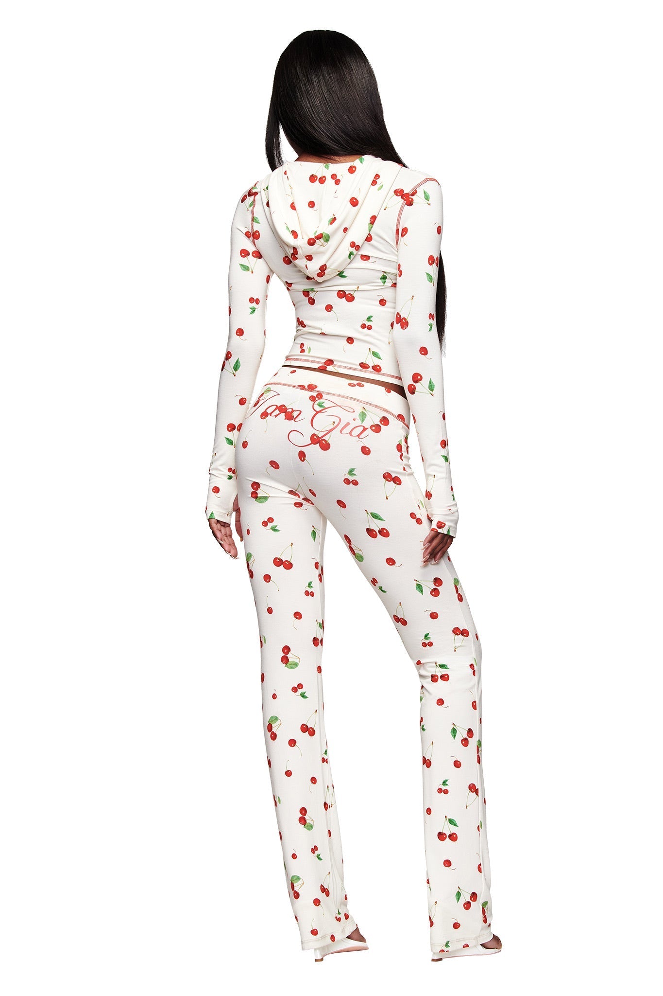 A person with long dark hair is shown from the back wearing the I.AM.GIA BLARE HOODIE - CHERRY ON TOP, which is white and features a red cherry print. The pants display cursive text and a playful cherry design on the back.