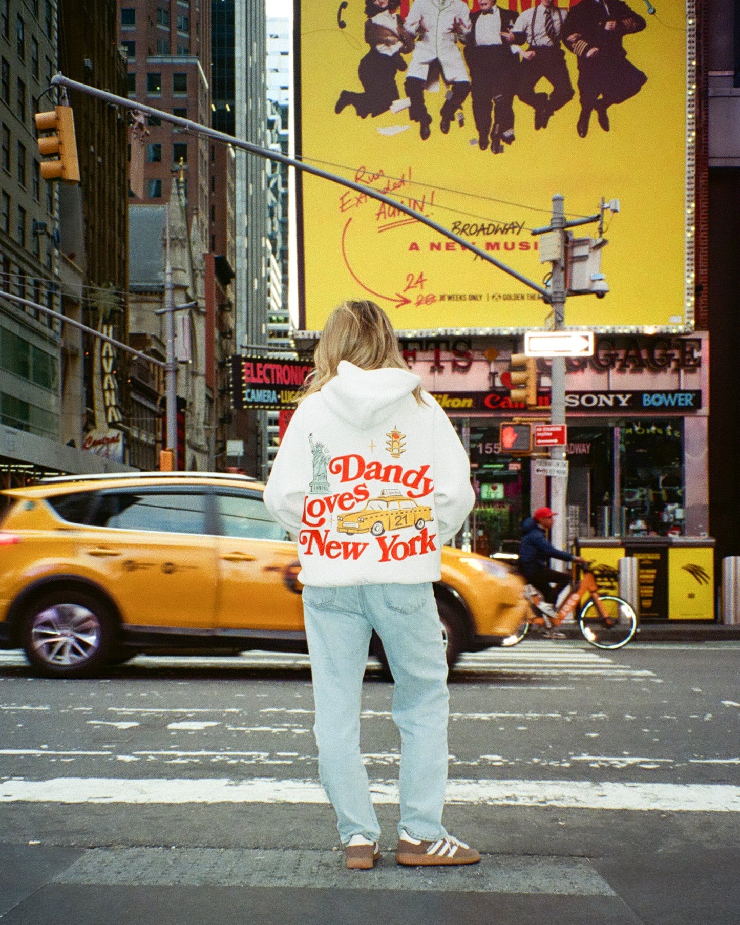 Person wearing a hoodie with text standing on a city street with a yellow taxi and large billboard in the background.