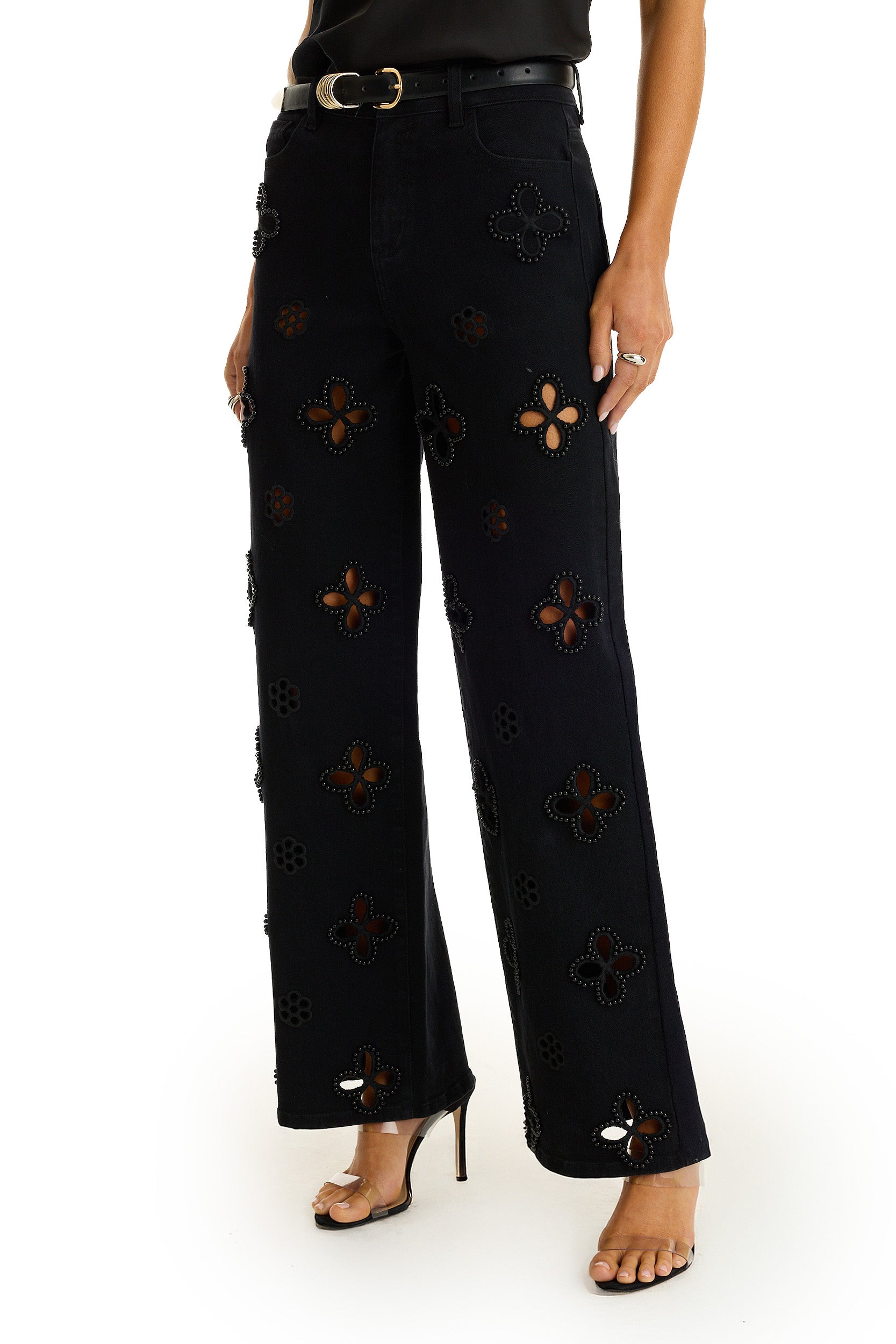 Wearing the Kaia Pearl High Rise Jean in black, featuring wide legs, cutout floral patterns, and pearl embroidery, paired with black heeled sandals and a tucked-in black top. The shot is cropped from the shoulders down.
