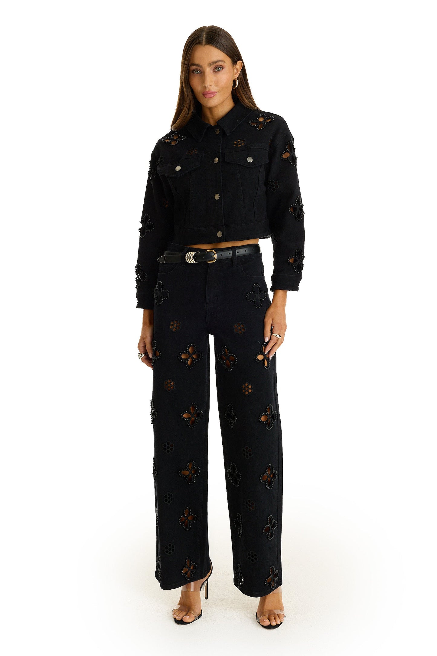 A woman stands facing forward in the Kaia Pearl High Rise Jean - Black, featuring cut-out floral patterns and pearl embroidery, paired with a matching black jacket, gold-buckle belt, and open-toe heels against a white background.