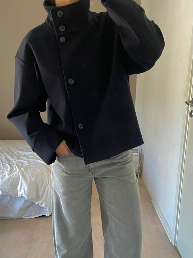 Black high-neck jacket with asymmetrical buttons and structured silhouette, featuring wide sleeves and a minimalist design for modern everyday styling.